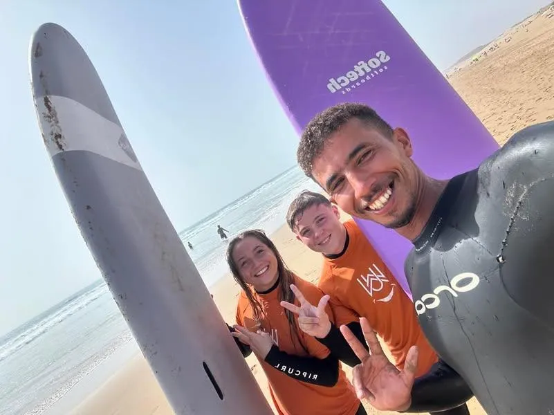 Two surf students in orange rashguards and coach Simo posing on the beach in Agadir