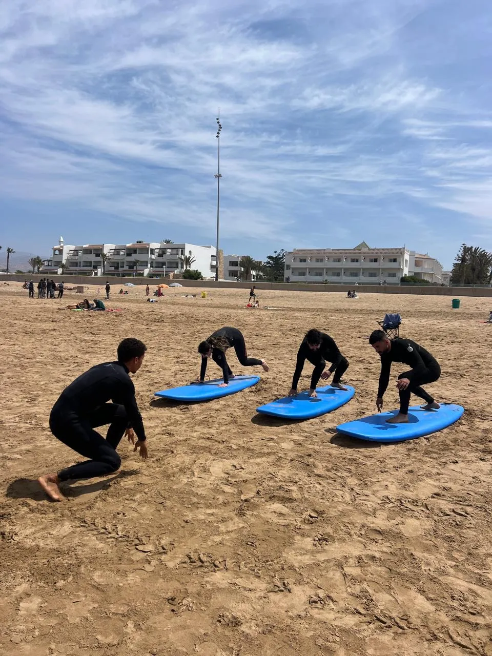 Group of four surf students practising the pop-up stance in unison on the sand at Agadir Beach with coach Simo at the front