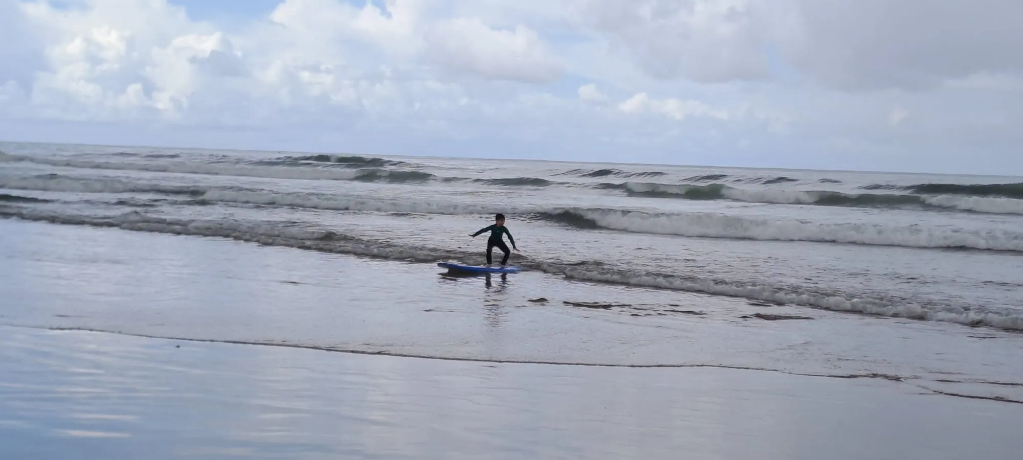Surf student standing on a blue board on a small wave in the Atlantic off Agadir, coach Simo not visible but watching from the shore