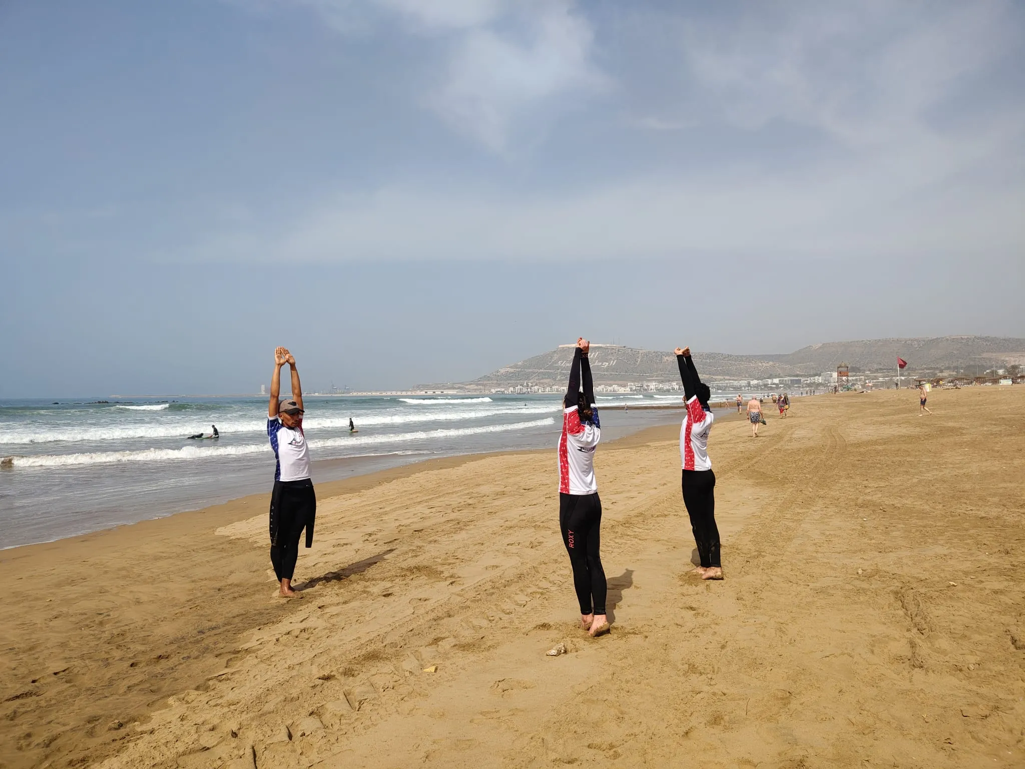 Three surf students stretching with arms above their heads during the pre-session warmup on Agadir Beach