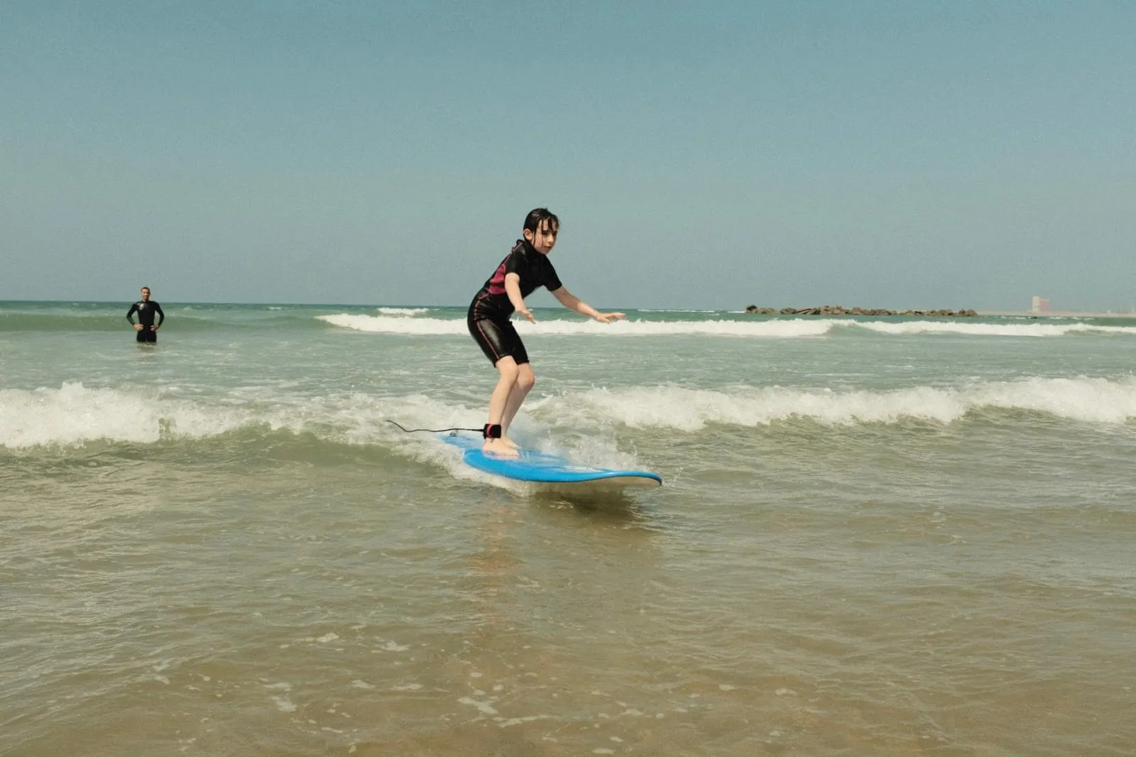 Young surf student standing on a blue surfboard catching a wave at Agadir Beach with coach Simo nearby in the water